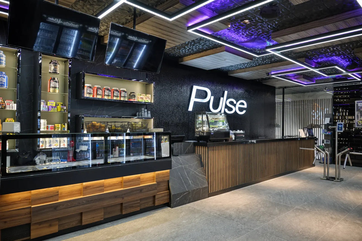 Reception and protein bar of a Pulse Fitness & Spa club, with the illuminated Pulse logo, supplement shelves and entry turnstiles