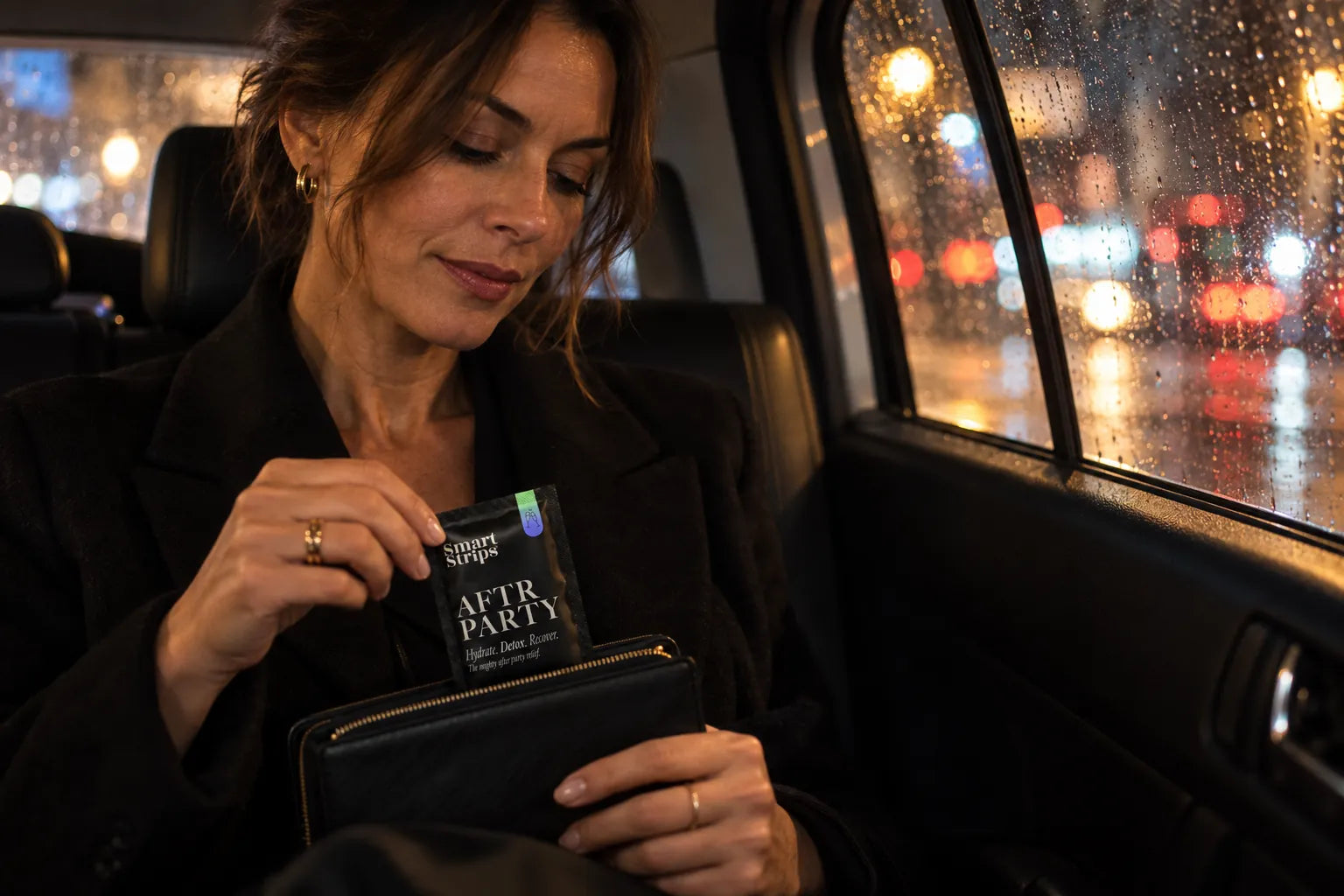 Woman in the back seat of a taxi at night holding a Smart Strips AFTR PARTY sachet, city lights on the rainy window